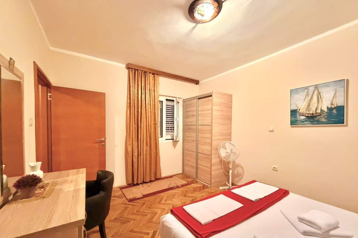 A hotel room with a wooden herringbone floor, a bed with white linens and a red accent, a desk with a chair, a door, a window with curtains, a fan, and a painting of sailboats on the wall.
