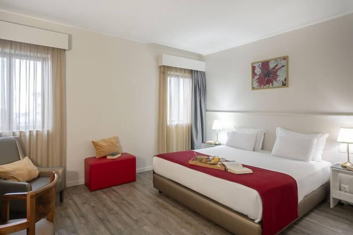 A hotel room with a bed, a chair, a small table, a lamp, and a painting on the wall.