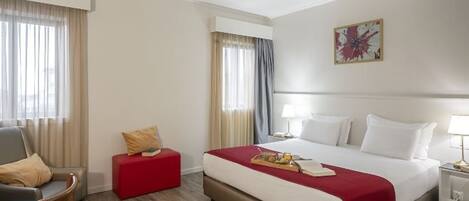 A hotel room with a bed, a chair, a small table, a lamp, and a painting on the wall.