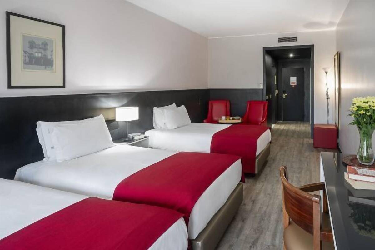 A hotel room with three beds, a desk, a chair, a lamp, and a framed picture on the wall.
