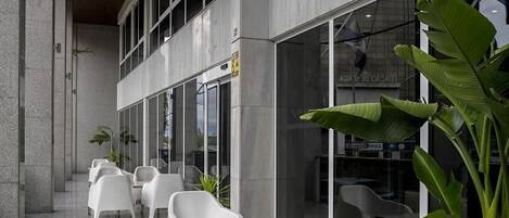 Un espacio de estar al aire libre con techo, sillas y mesas blancas, ventanas amplias y plantas en macetas.