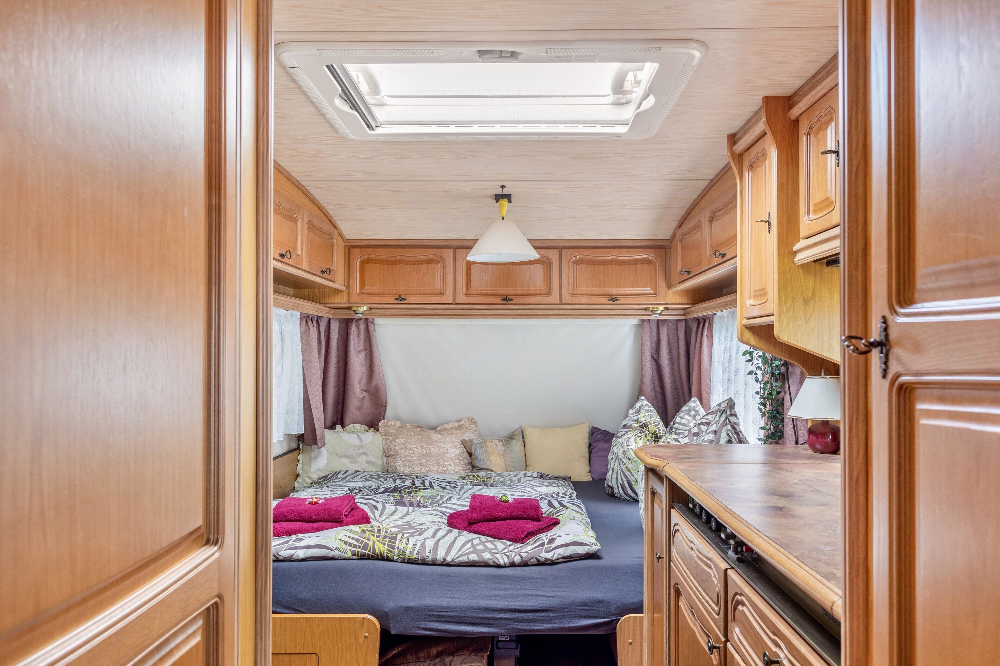 Dormitorio compacto de autocaravana con cama, armarios de madera, kitchenette y tragaluz.
