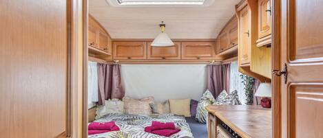 Dormitorio compacto de autocaravana con cama, armarios de madera, kitchenette y tragaluz.