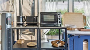 Une petite cuisine équipée d’une machine à café, d’un grille-pain et d’un four à micro-ondes, le tout posé sur une étagère en métal, ainsi qu’une planche à découper en bois et un panier sur une table.