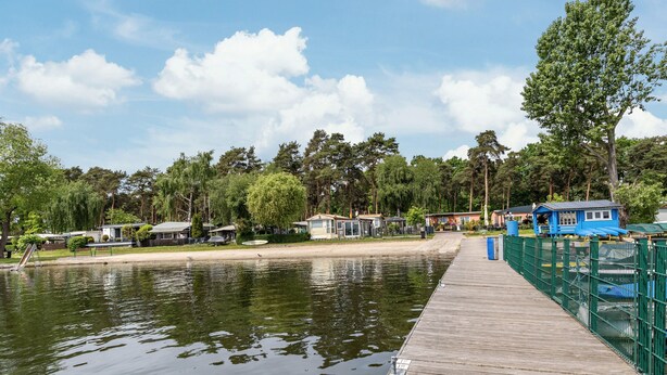Une jetée en bois s’étendant sur une étendue d’eau calme, entourée d’arbres et de petits bâtiments.
