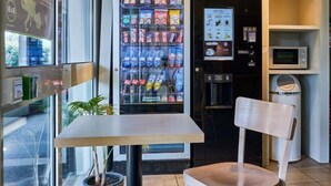 Máquina expendedora con snacks, microondas y cubo de basura en una habitación con una mesa y una silla.