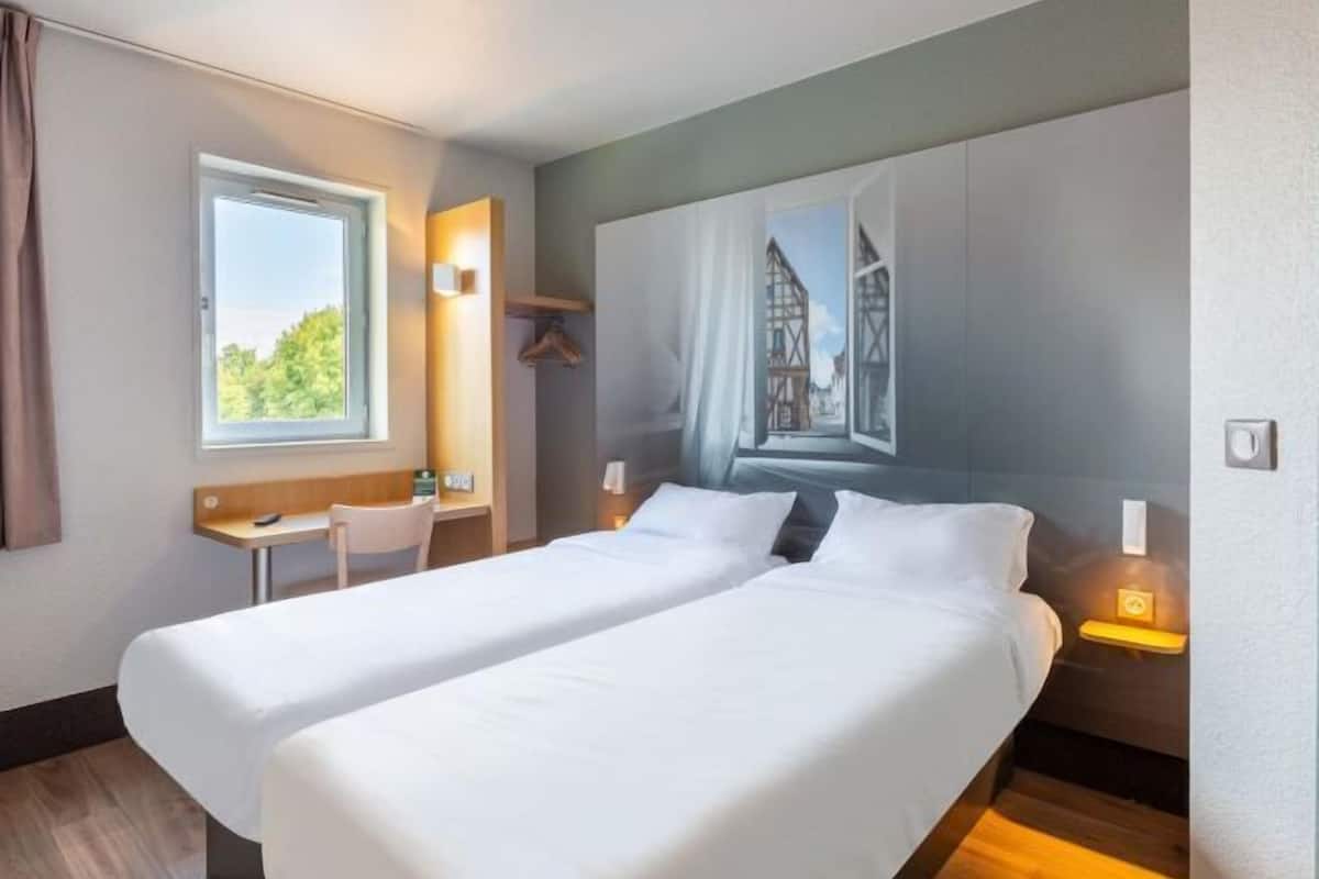 Habitación de hotel con dos camas, un escritorio, una silla y una ventana con vistas a zonas verdes.