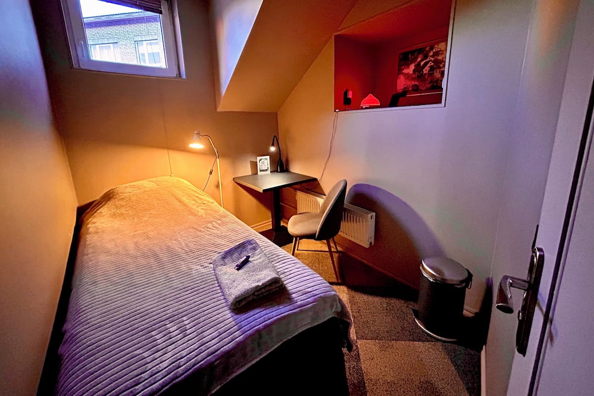 A small, dimly lit room with a single bed, a desk, and a chair. There is a window providing natural light, a radiator, and a door.