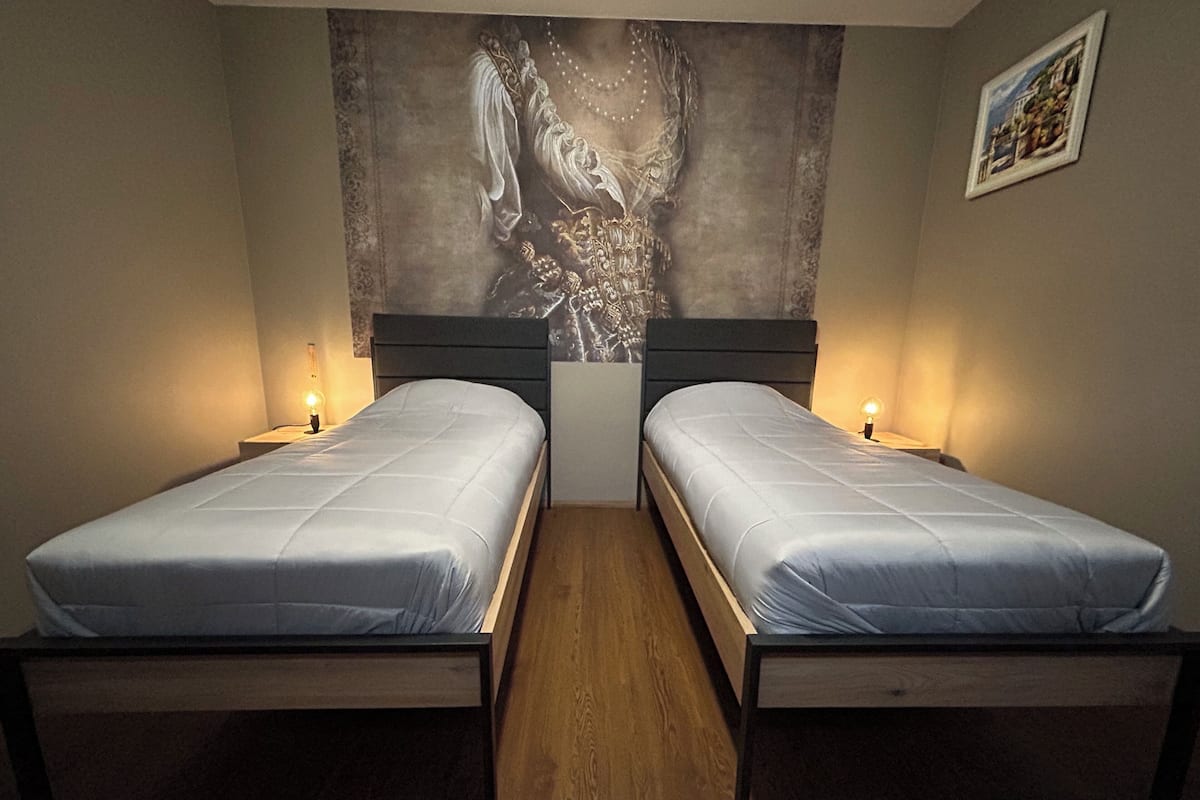 A room with two single beds, each with a wooden frame and a white mattress. There are bedside tables with lamps on either side of the beds. The room has a large, dark-colored tapestry hanging on the wall above the beds.