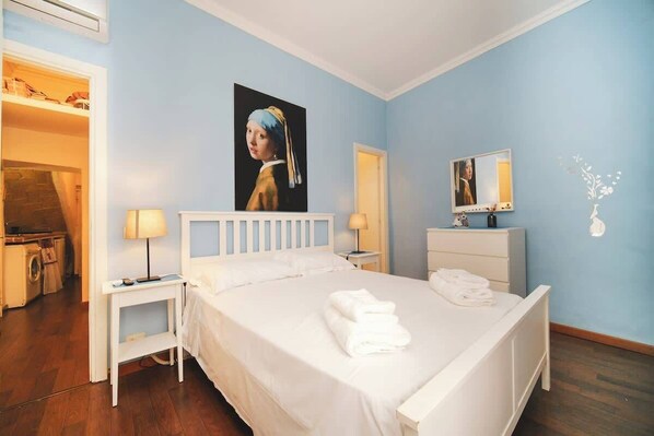 A bedroom with a bed, bedside tables, a mirror, a painting, and a bathroom visible through an open door.