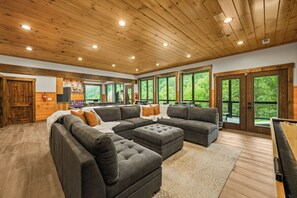 A spacious living area with a sectional sofa, a pool table, and large windows offering a view of the greenery outside.