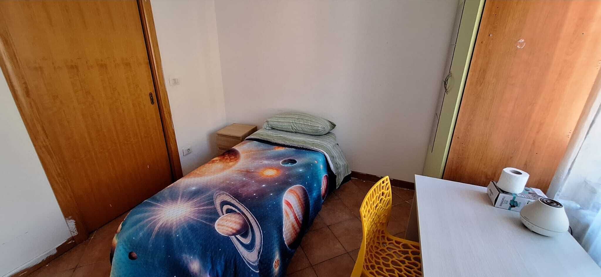 A small room with a bed covered in a space-themed bedspread, a wooden door, a small table with a chair, and a window with curtains.