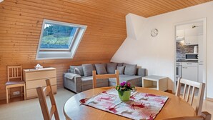 A cozy room with a wooden ceiling, a dining table with a floral table runner, a grey sofa, and a small kitchen area. - 'Elzblick' Apartment with Shared Pool, Garden, and Wi-Fi (Elzach)