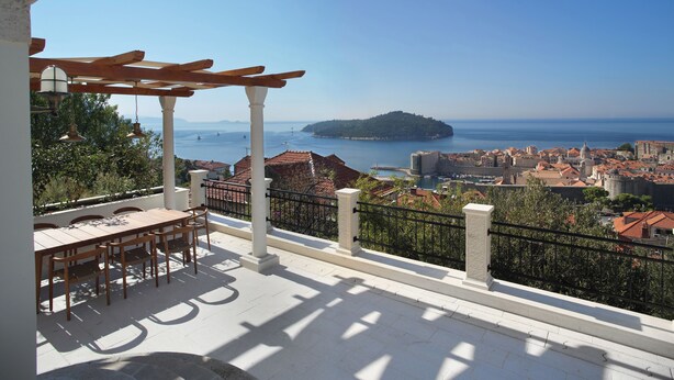 Eine Terrasse mit einer Pergola, einem Esstisch und Stühlen mit Blick auf eine Küstenstadt, historische Gebäude und den Meerblick.