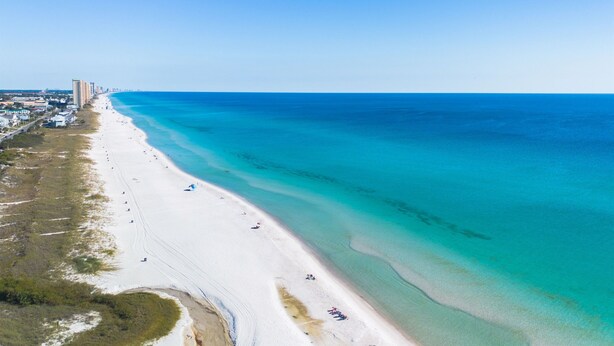 Vista aérea de una playa con una extensa franja de arena blanca, agua turquesa cristalina y un horizonte urbano distante con edificios.