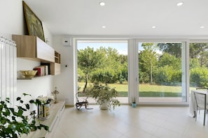 A modern interior with a large sliding glass door leading to a garden, a white tiled floor, and a bookshelf with decorative items. - Apartment 1 with Lake View (Alvignano)