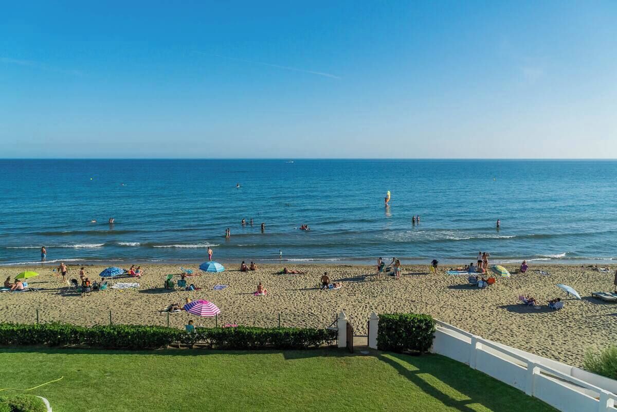 Ein Strand mit badenden und sonnenden Personen, ein klarer Himmel und ein Zaun im Vordergrund.