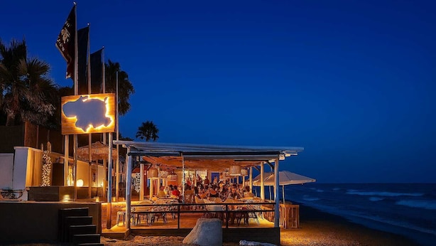 A beachfront restaurant with a covered seating area, a large illuminated sign, and flags. - Beach House in front the sea (Marbella)