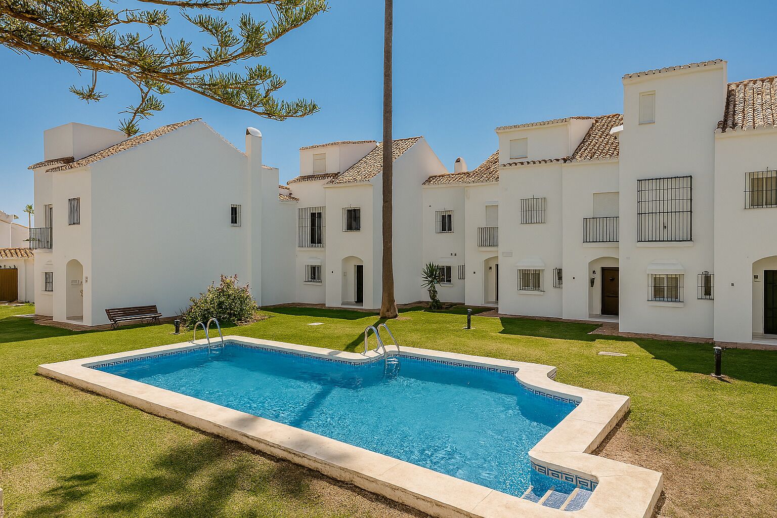 Une piscine entourée de bâtiments blancs dotés de balcons, une pelouse verdoyante et un ciel bleu azur.