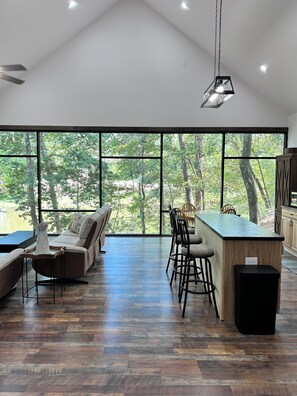 Ruime kamer met groot raam, houten vloer, een bank, een bar en een plafondventilator.