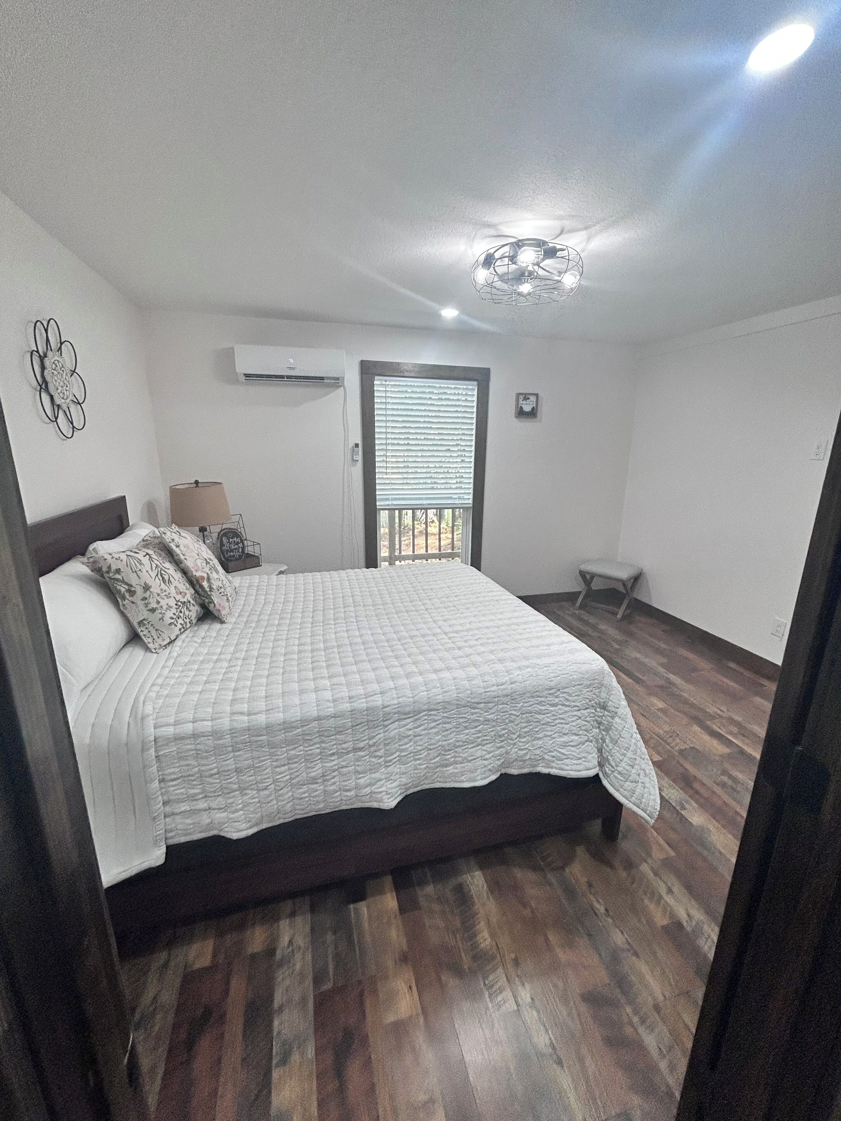 Un dormitorio con cama, ventana con persianas, aire acondicionado y una mesita de noche con lámpara.