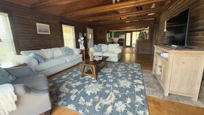 A log cabin interior with a sofa, armchairs, a coffee table, a TV, and a kitchen area. - Lakeside Log Cabin Retreat- Close to Beach + Fishing + Boat Parking (Corpus Christi)