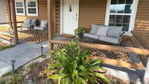 A wooden house with a porch, two windows, a door, and a seating area with a sofa and chairs. - Lakeside Log Cabin Retreat- Close to Beach + Fishing + Boat Parking (Corpus Christi)