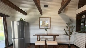 A kitchen with a refrigerator, a table with two chairs, a potted plant, and a framed picture on the wall. - Lakeside Log Cabin Retreat- Close to Beach + Fishing + Boat Parking (Corpus Christi)
