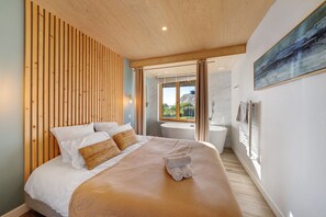 A modern bedroom with a wooden slat headboard, a large bed with white linens, a view of the outdoors through a glass door, and a painting on the wall.