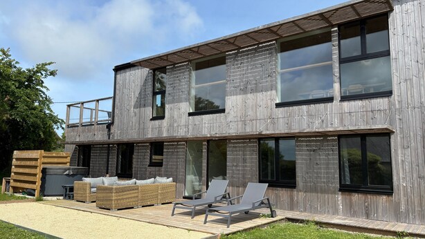 Un bâtiment moderne à deux étages, doté de grandes fenêtres, d’une terrasse en bois avec des meubles d’extérieur et d’un balcon.