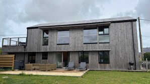 Une maison moderne à deux étages, construite en bois, dotée de grandes fenêtres, d’une terrasse et d’un espace salon extérieur.
