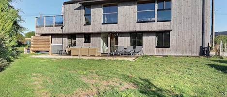 A two-story wooden house with a flat roof, large windows, and a covered patio area with outdoor furniture.