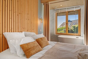 A bedroom with a wooden headboard, a bed with white and brown pillows, a window with a view of a building and trees, and a wall-mounted light fixture. - Villa Grande Est, Private Spa, Ideal for Family Holidays (Plouneour-Trez)
