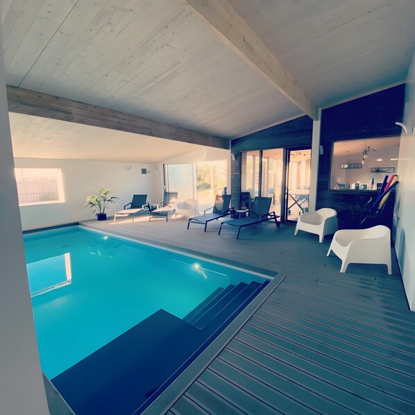 A modern indoor swimming pool with lounge chairs and a view of the outdoors.