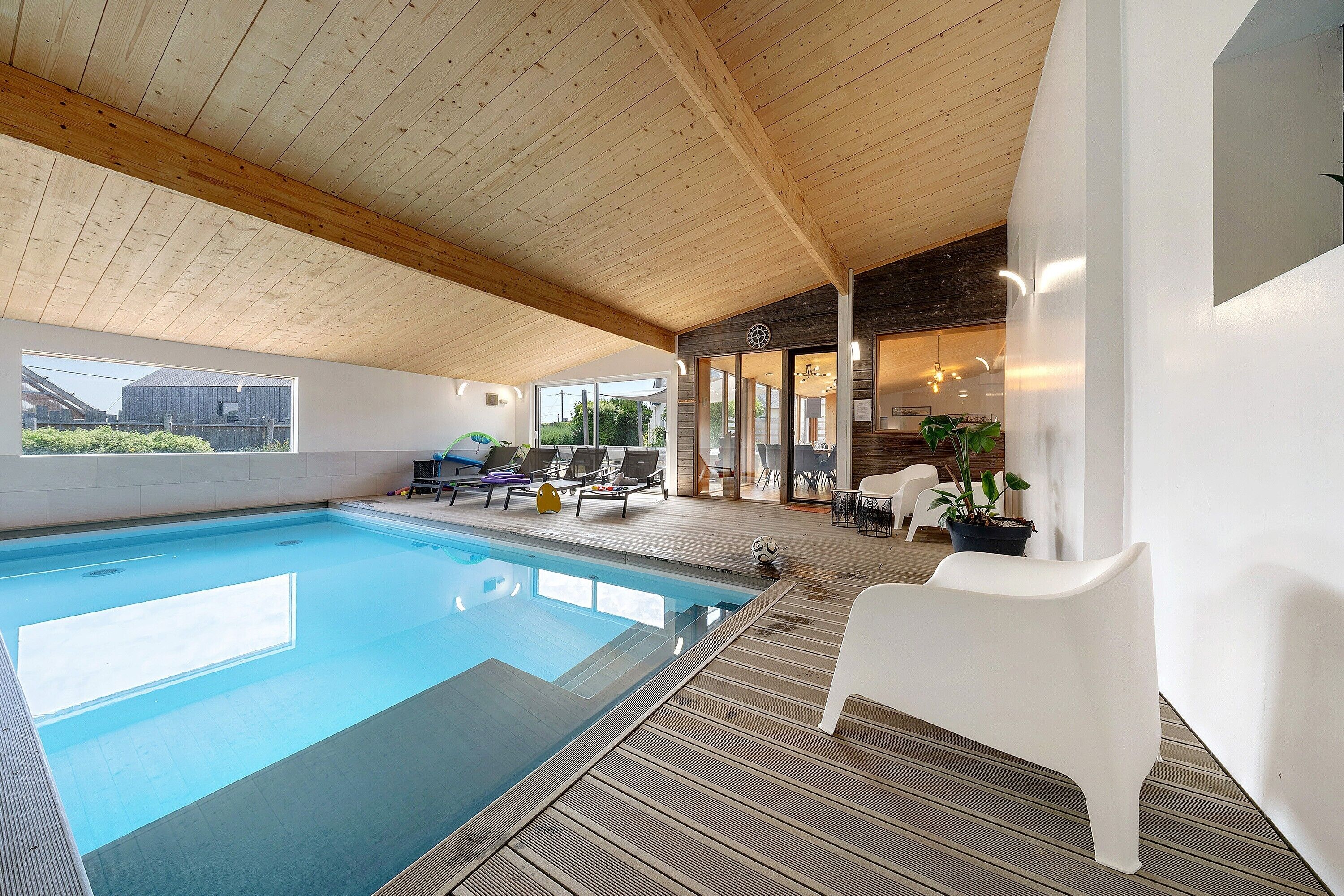 Een binnenzwembad met een houten plafond, ligbedden en uitzicht op buiten.
