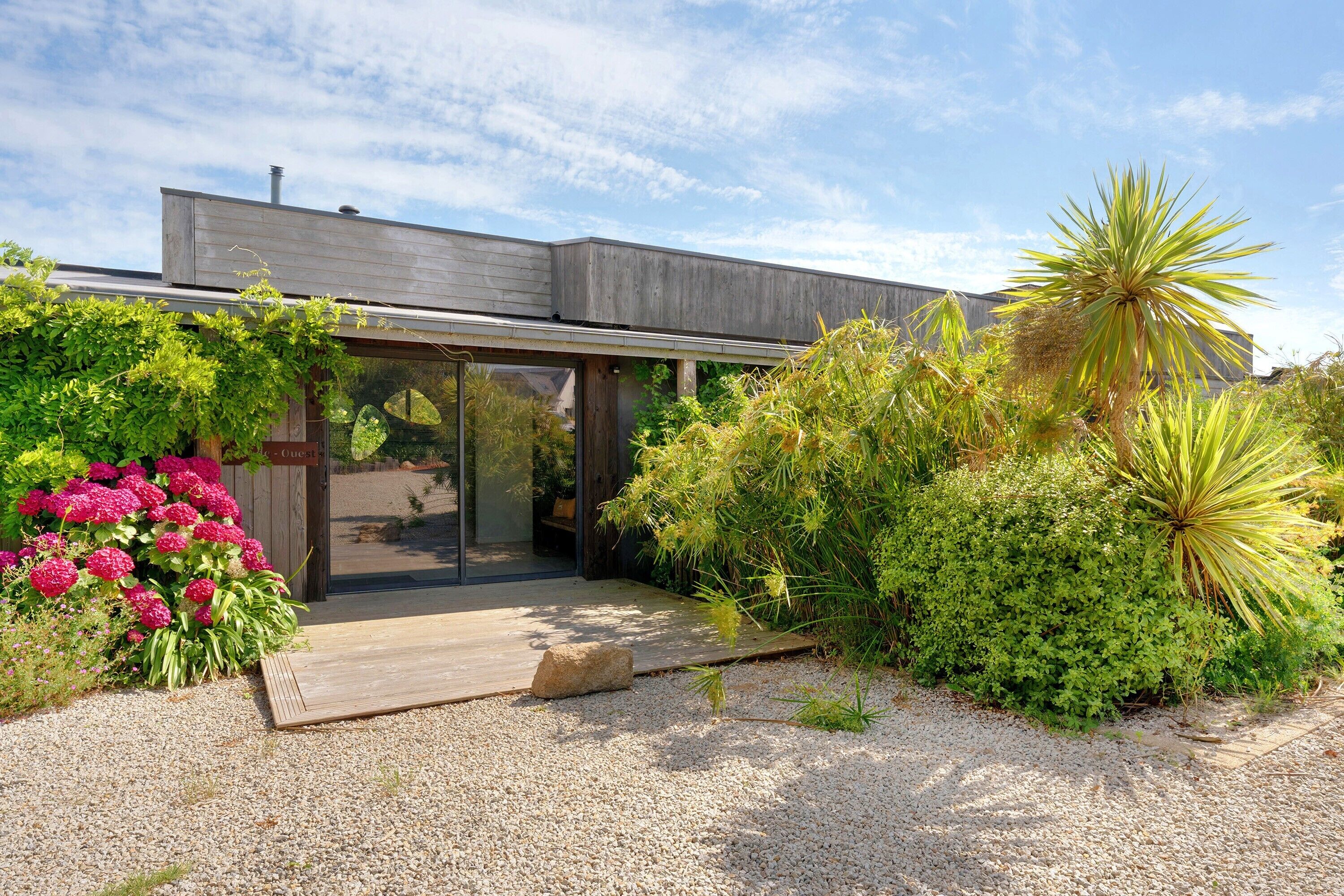 Een modern huis met een houten terras, een glazen entree en een tuin met diverse beplanting.
