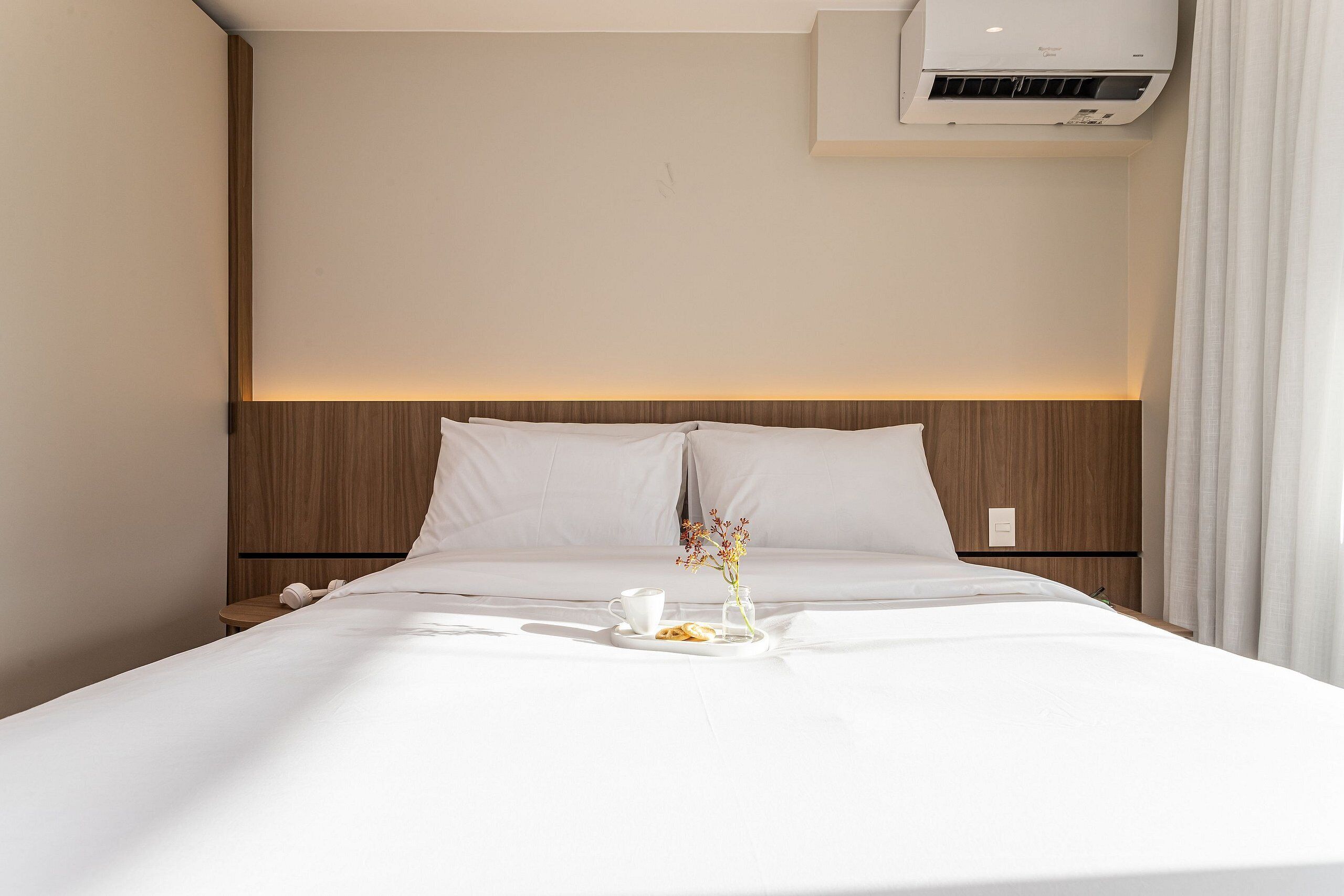 Ein Hotelzimmer mit einem ordentlich bezogenen Bett, einem Nachttisch mit Tasse und einem Tablett mit Speisen sowie einer wandmontierten Klimaanlage.
