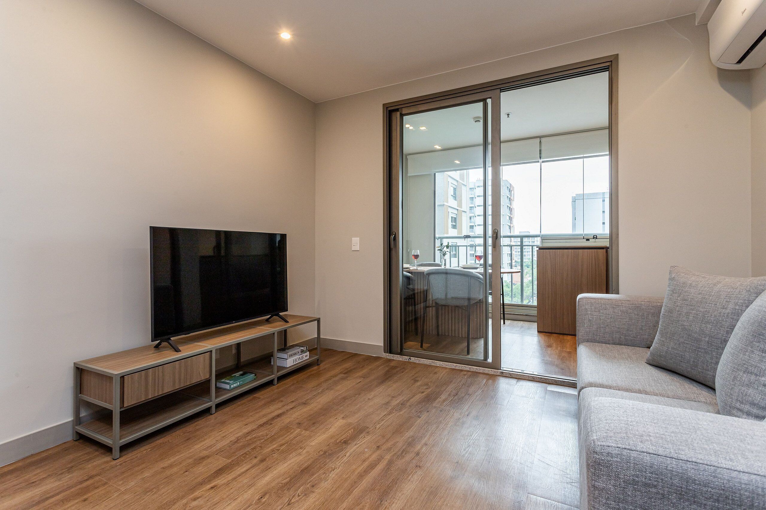 A modern living room with a flat-screen TV on a wooden TV stand, a grey sofa, and a sliding glass door leading to a balcony.