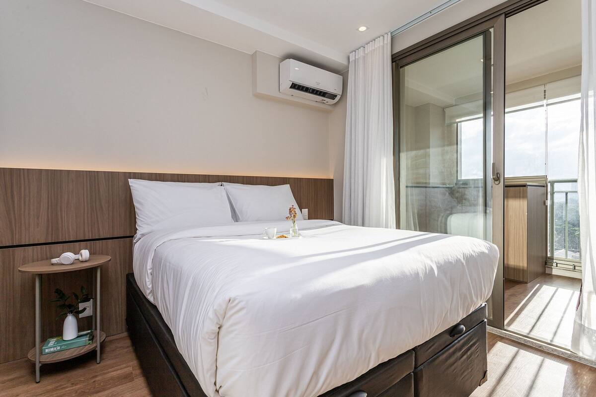 A hotel room with a bed, bedside table, air conditioning unit, and a view of the outside.