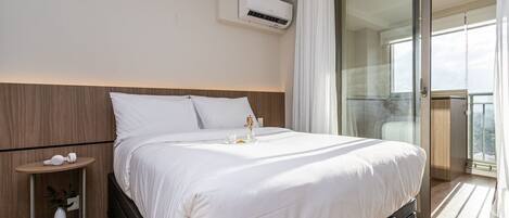 A hotel room with a bed, bedside table, air conditioning unit, and a view of the outside.