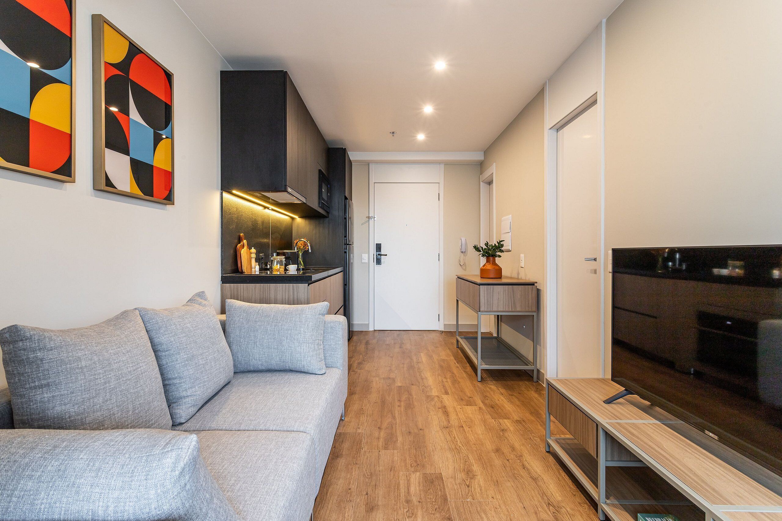 A modern living room with a grey sofa, wooden flooring, a kitchen area with a TV, and abstract wall art.