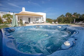 Un bain à remous avec un bord carrelé, un bâtiment blanc avec un toit en tuiles et des sièges d’extérieur.