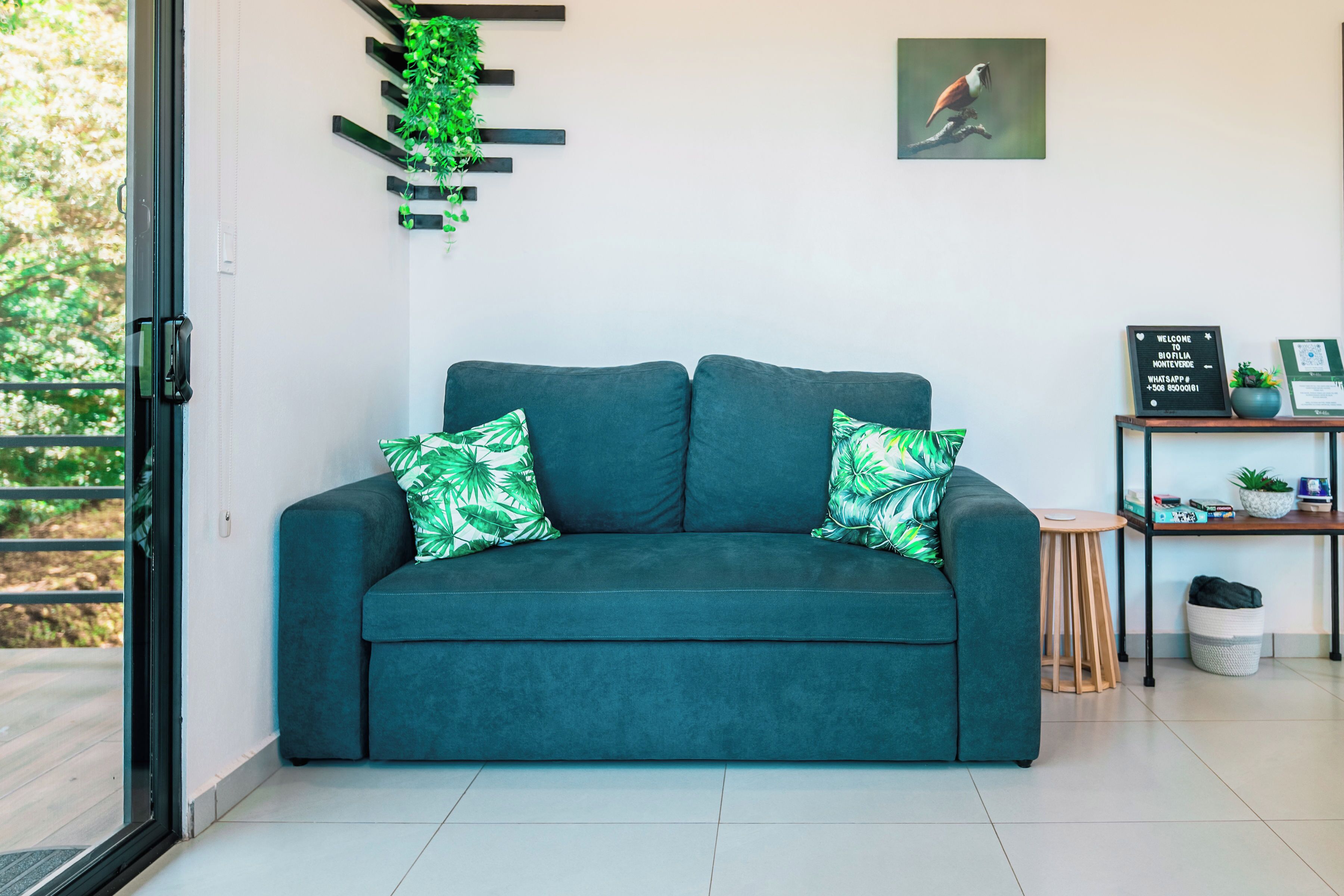 Un sofá azul verdoso con dos almohadones, una planta montada en la pared, un cuadro enmarcado, una mesita con una planta y un letrero de bienvenida.