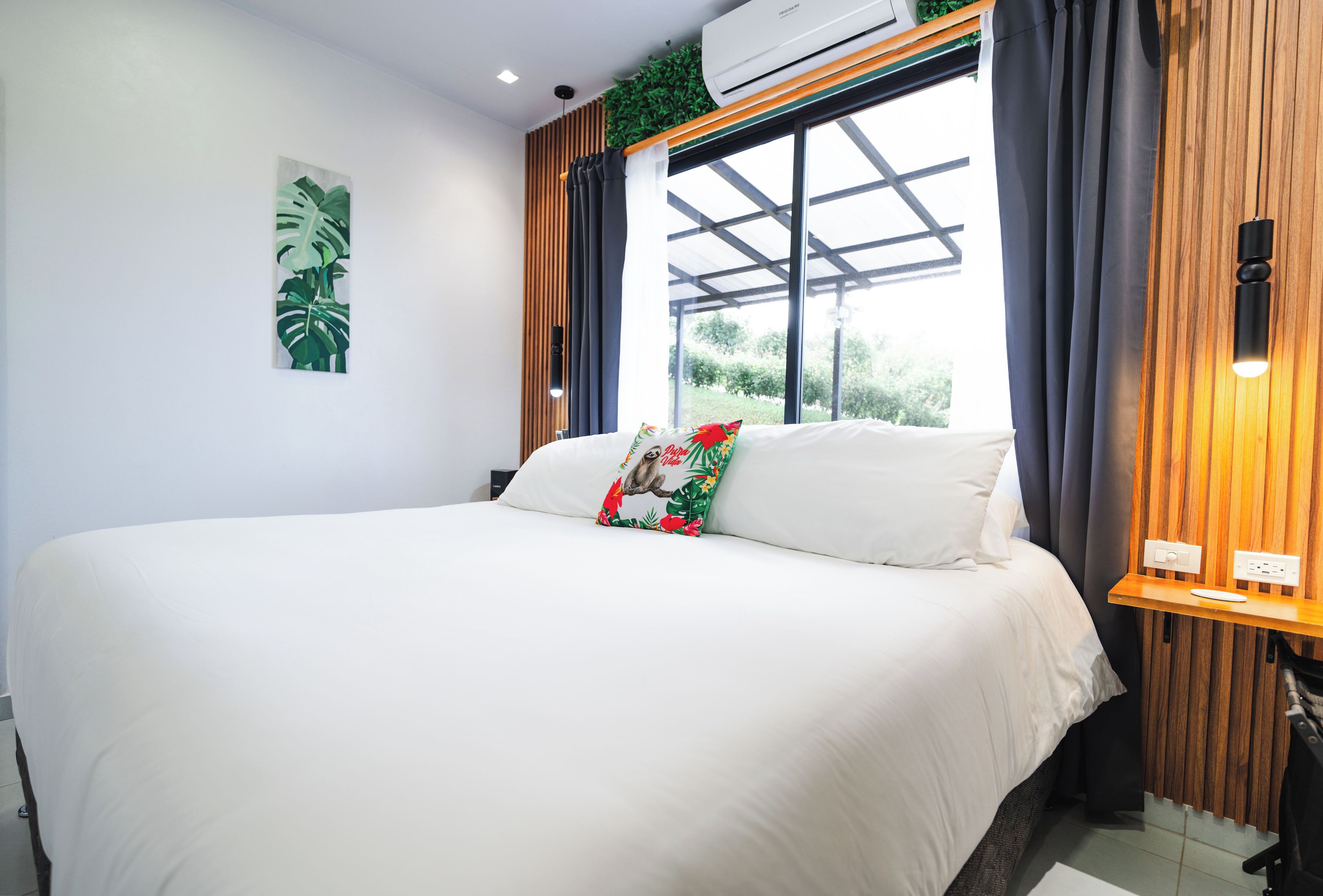 Ein modernes Schlafzimmer mit einem großen Bett, einem Fenster mit Vorhängen, einer Wand-Klimaanlage und einem kleinen Nachttisch mit Lampe.