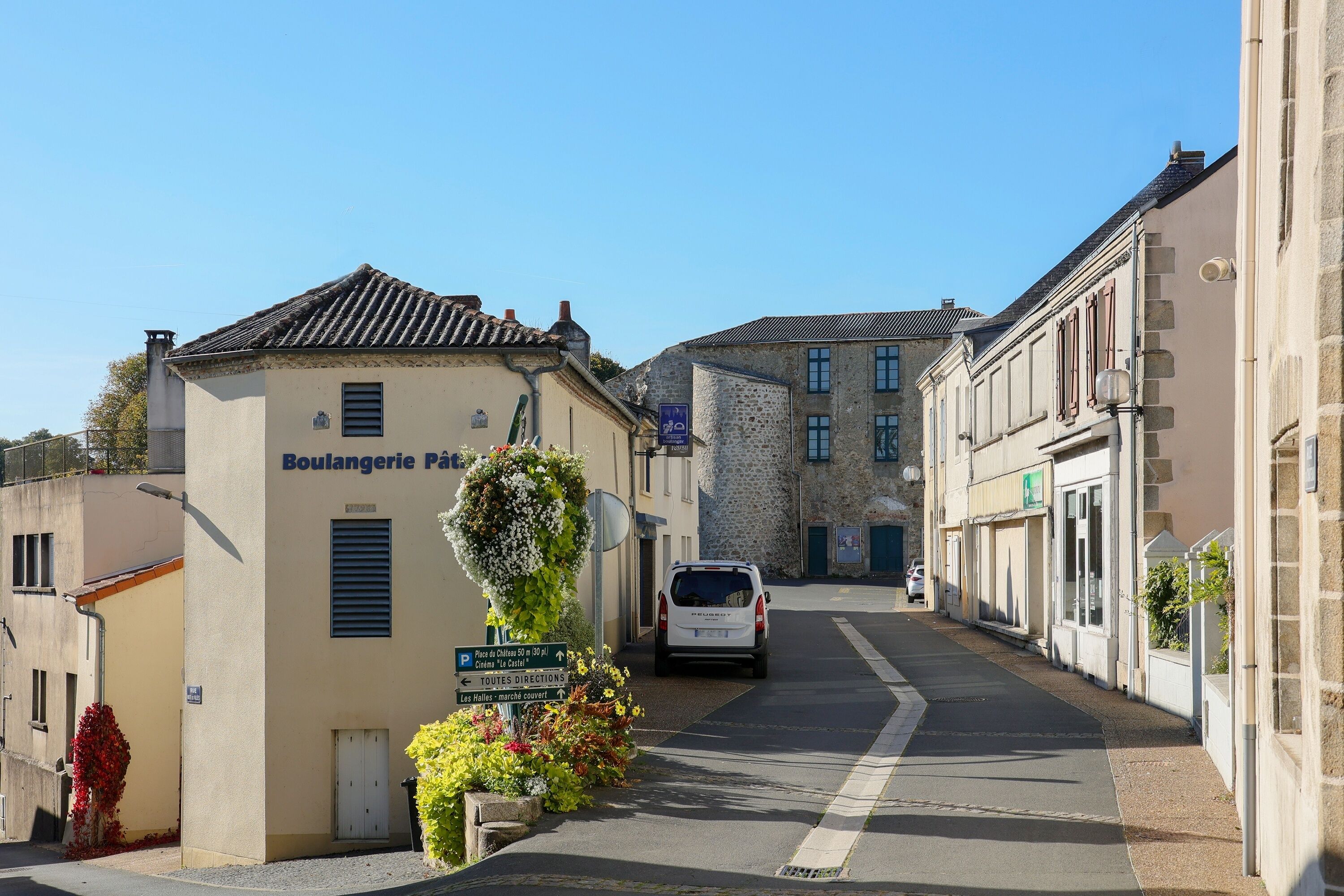 Une rue étroite, bordée de bâtiments des deux côtés, avec une voiture garée et une enseigne indiquant une « boulangerie-pâtisserie ».