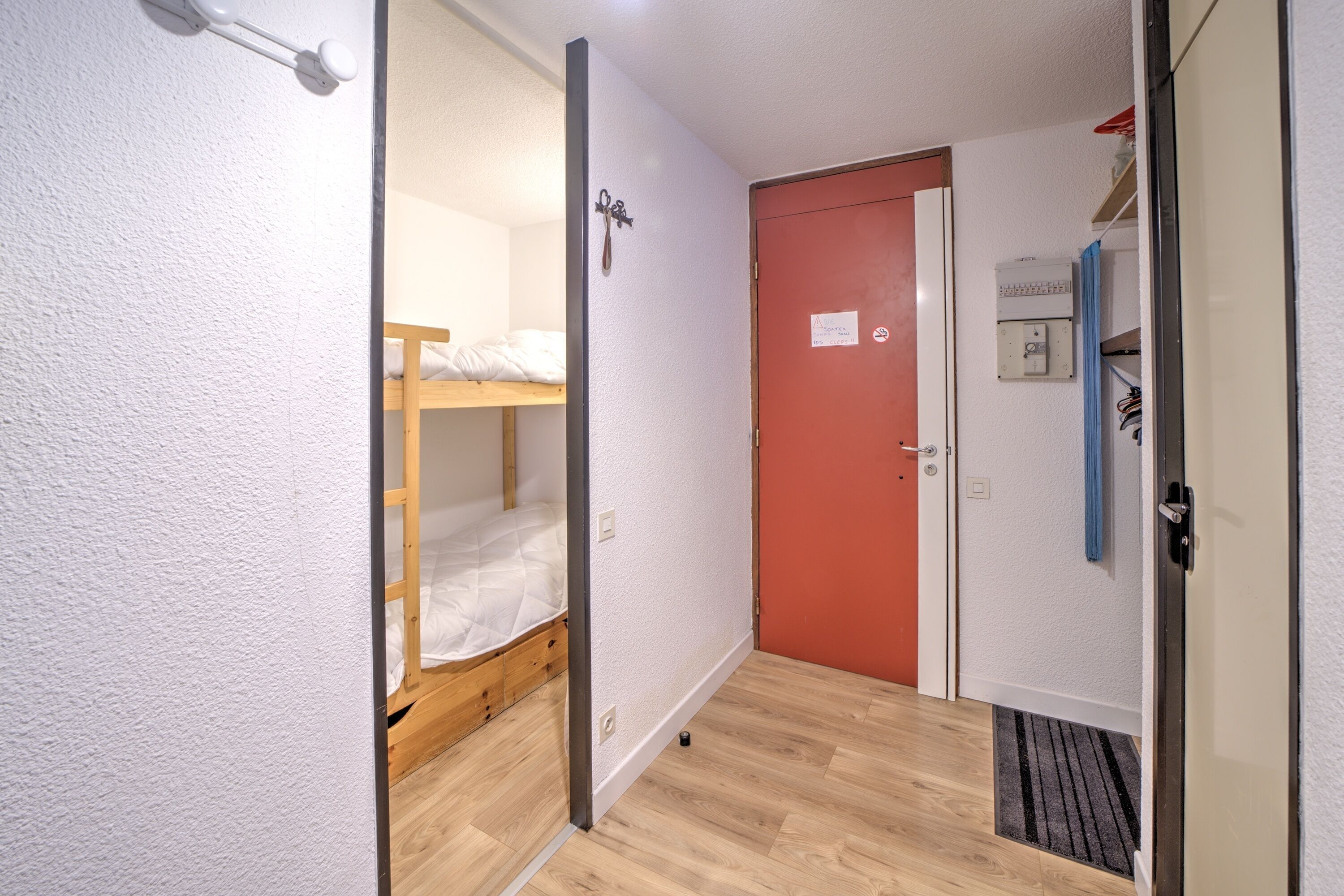 Un pasillo estrecho con una cama doble de madera, una puerta roja y una pared blanca con un interruptor de luz.