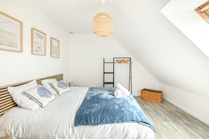 A bedroom with a sloped ceiling, a bed with white bedding and blue accents, a wooden headboard, a hanging light fixture, and a framed picture on the wall. - House of Travels - Proche Plage (Manvieux)