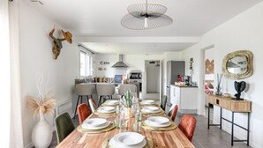 A modern dining area with a wooden table set for six, surrounded by chairs. A large pendant light hangs above, and the room features a white wall with decorative items.