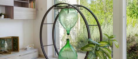 Una habitación con un amplio ventanal, un jarrón verde sobre un pedestal, una planta en maceta y una estantería.