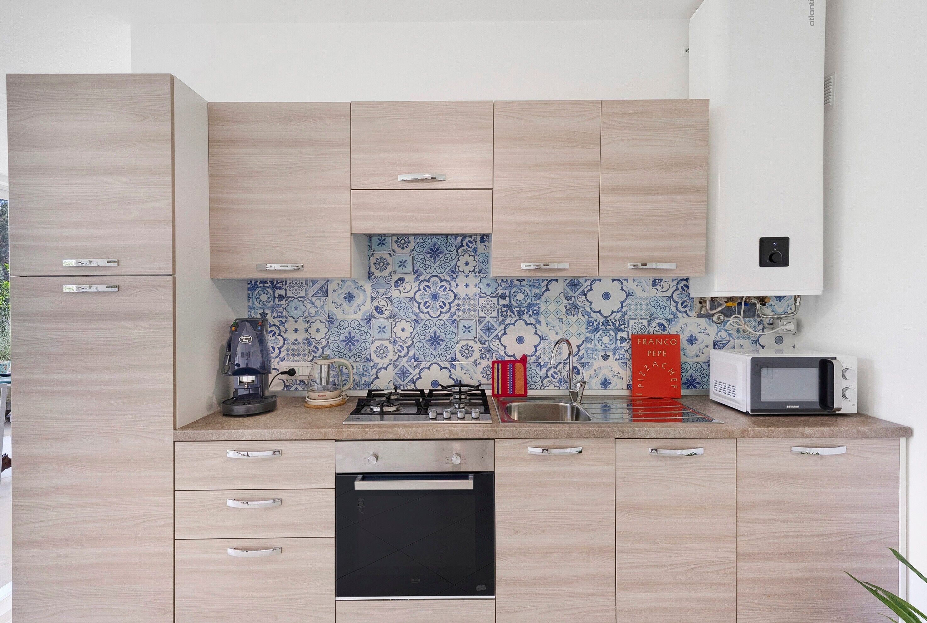 Una cucina moderna con mobili chiari in legno, lavastoviglie nera, piano cottura a gas, macchina del caffè, forno a microonde e bollitore rosso.